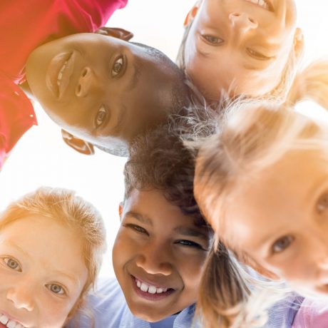 Portrait of happy kids in circle looking down and embracing. Group of five multiethnic friends outdoor looking at camera and smiling. Closeup face of smiling children looking down at the camera together at park.